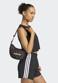 Torba na ramię z materiału w panterkę z białym logo "adidas", o zaokrąglonym kształcie, z regulowanym paskiem, zestawiona z czarnym krótki topem i szortami.