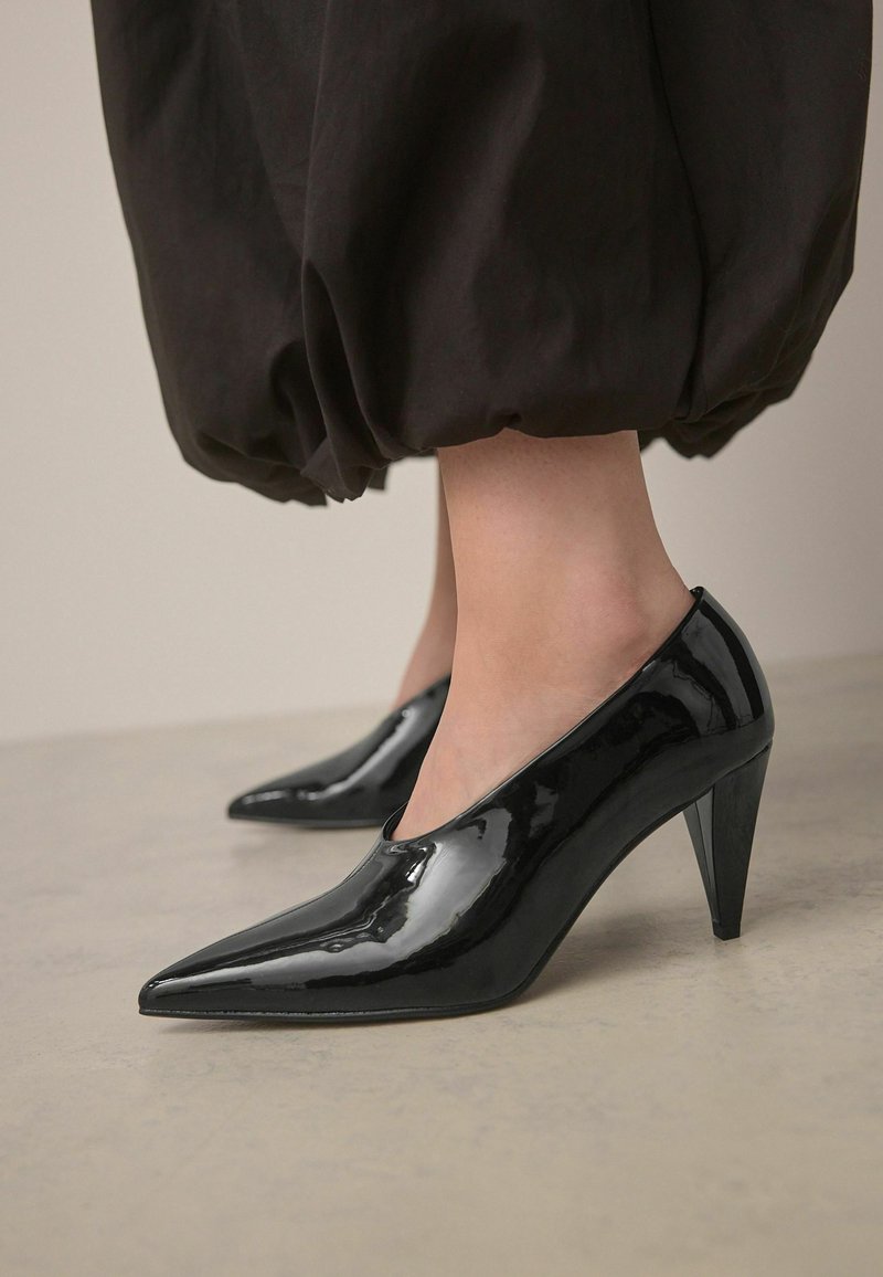 Zapatos de tacón alto de charol negro con puntera afilada y un delgado tacón de aguja, que presentan un acabado suave y brillante.