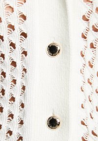 Cardigan bianco lavorato a maglia con un motivo in pizzo su un lato, dotato di un grande bottone dorato lucido per la chiusura.