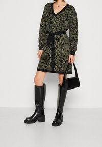 Abito in maglia con stampa a serpente verde e nera, scollo a V, maniche lunghe, vita con cintura, abbinato a stivali neri sopra il ginocchio e una borsa nera.