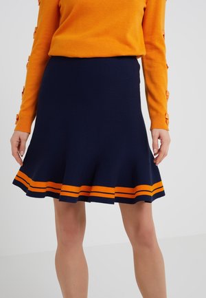 Frau trägt einen marineblauen ausgestellten Rock mit zwei orangefarbenen Streifen nahe dem Saum und ein oranges Langarmoberteil mit seitlichen Knopfdetails.