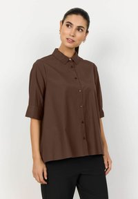 Chemise brune à manches courtes, boutonnée, fabriquée en tissu lisse avec un col et une coupe ample, présentant un ourlet droit et des boutons assortis.