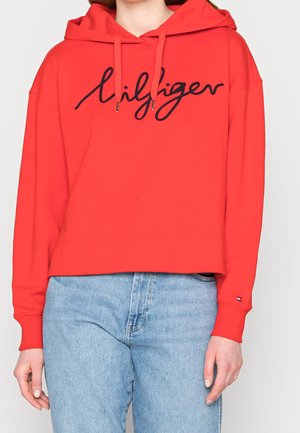 Sudadera roja recortada con texto en cursiva negra "Hilfiger", cordones y vaqueros azul claro en una persona de pie contra un fondo liso.