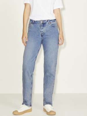 Person iført lyseblå jeans med lige ben, hvid t-shirt og hvide sneakers med beige såler, stående mod en ensfarvet baggrund.