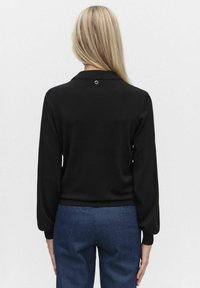 Musta pitkähihaine paita, jossa on pyöreä kaulus, ribatulla hihansuulla ja pieni hopeinen yksityiskohta takana. Sileä pinta, tiukka malli.