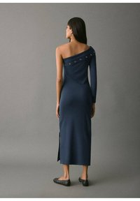 Abito blu navy a spalla unica con silhouette aderente, caratterizzato da accenti metallici lungo il collo e una spacco laterale per una maggiore libertà di movimento.
