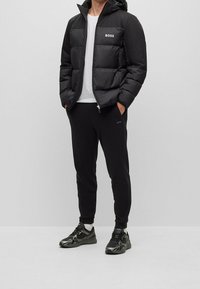 Svart pufferjacka med huva och logotyp på bröstet, tillsammans med svarta joggers och sportskor. Slät tyg, dragkedjestängning och elastiska mudd.