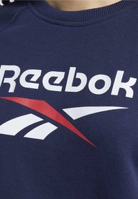 Mörkblå sweatshirt med rund halsringning som har stor vit "Reebok" text och en röd accentdesign. Texturen är mjuk och jämn.