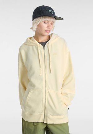 Geel ritsvest met een buidelzak, ribgebreide manchetten en een zachte textuur. Gekocht met een grijs shirt en groene broek; aangevuld met een donkere pet.