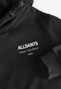 Sudadera negra con una textura suave, que cuenta con una capucha con cordón, puños de canalé y un logotipo blanco de la marca con texto en japonés e inglés.