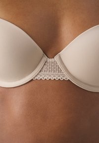 Beige beha met gladde, gevormde cups en kanten rand. Het materiaal is zacht met een lichte glans en heeft een decoratieve kanten inzet aan de achterkant.