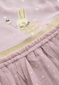 Tissu lilas avec une texture à côtes présentant un motif de lapin. Bordure dorée et volan en tulle en bas avec de petits pois dorés.