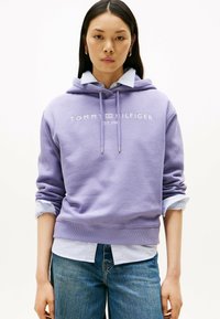 Paarse katoenen hoodie met logo, trekkoord en geribbelde mouwen, gelaagd over een lichtblauw gestreept shirt, gecombineerd met een spijkerbroek.