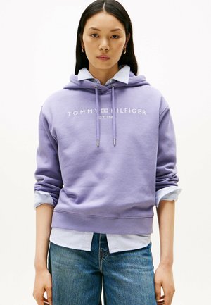 Haut en coton violet avec logo, cordons de serrage et poignets côtelés, superposé à une chemise rayée bleu clair, associé à un jean en denim.