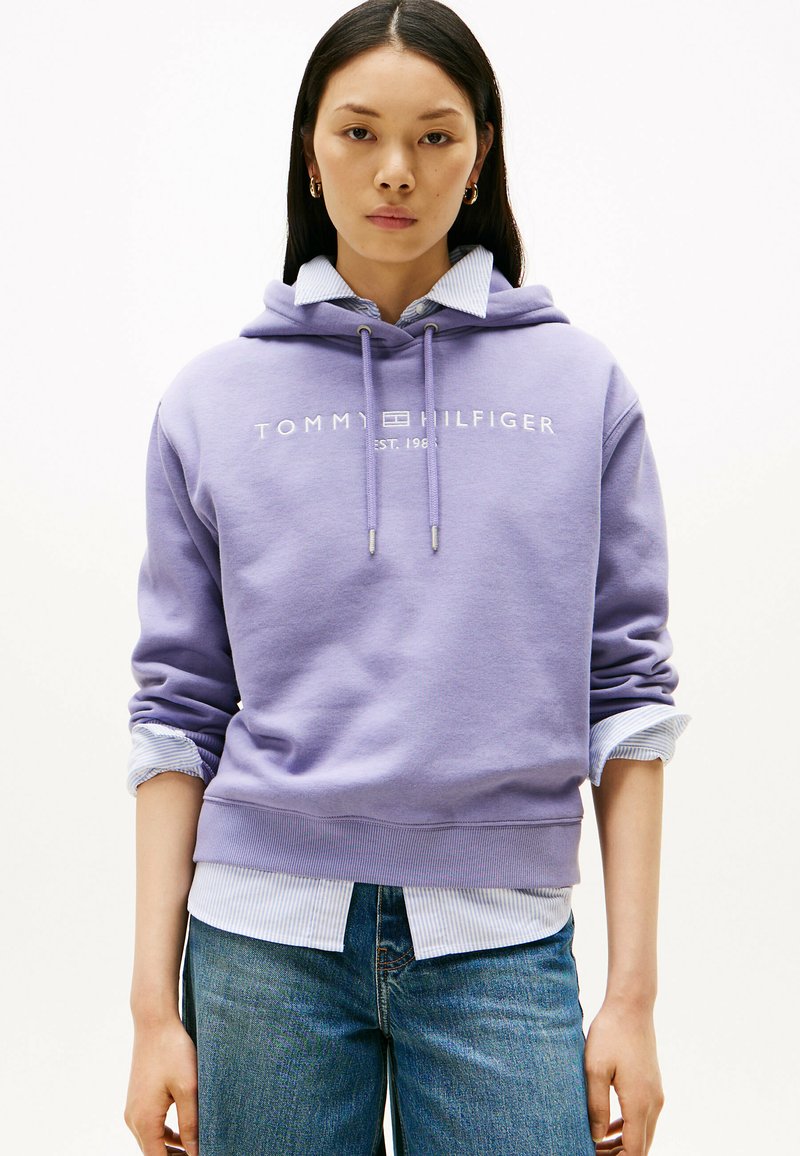 Paarse katoenen hoodie met logo, trekkoord en geribbelde mouwen, gelaagd over een lichtblauw gestreept shirt, gecombineerd met een spijkerbroek.
