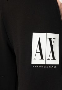 Svart tyg med vit rektangulär logotyp som visar stort "A|X" och mindre "Armani Exchange" text nedan.
