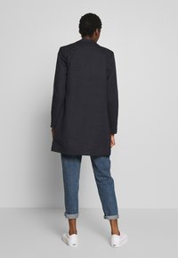 Længere mørkeblå frakke med krave, med en lige pasform og sidelommer, båret over opkrøllede blå jeans og hvide sneakers.
