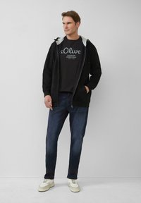 Czarna bluza z kapturem na zamek z szarym wykończeniem, czarna koszulka z białym napisem, ciemnoniebieskie dżinsy i kremowe trampki. Casualowy strój.