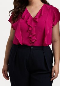 Fuchsia-blus med volangkrage och korta ärmar, i kombination med mörka högmidjade byxor. Slät textur och figursydd design.