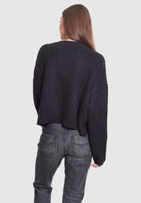 Maglione nero a maglia con vestibilità ampia e polsini a costine, abbinato a jeans grigi con texture. La vista posteriore rivela una lunghezza cropped.