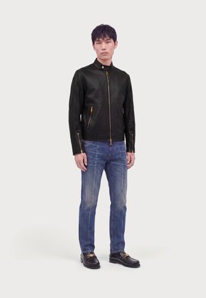 Jeune homme debout portant une veste en cuir noire zippée, un jean bleu et des mocassins noirs sur un fond blanc uni.