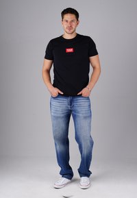Schwarzes Baumwoll-T-Shirt mit rotem rechteckigen Logo auf der Brust, kombiniert mit hellblauen Denim-Jeans und weißen Sneakers. Das Model steht mit den Händen in den Taschen.