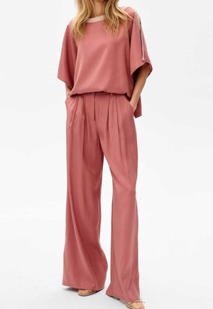 Femme portant un pantalon large ample couleur rose poussière et un haut ample à manches courtes assorti avec un bord blanc à l'encolure et aux manches.
