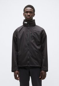 Chaqueta impermeable negra con cuello alto, cremallera frontal y bolsillo en el pecho con cremallera; incluye el logo de la marca en el cuello. Textura lisa.