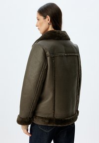 Oliwkowa kurtka z owczej wełny z szerokim kołnierzem, teksturowanym wykończeniem ze skóry i miękką podszewką. Posiada wyraźnie zarysowane ramiona i ściągacze przy mankietach.