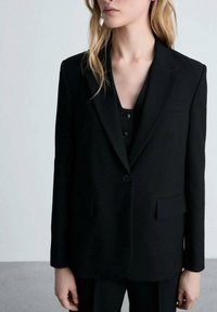 Černý oblekový blazer s klopami, jedním knoflíkem, dvěma předními kapsami a hladkým povrchem. Nesený přes sladěný vrstvený outfit.