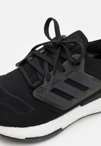 Czarne buty do biegania Adidas z dzianinową cholewką, grubą białą amortyzującą podeszwą oraz czarną plastikową klatką boczną z trzema paskami i związanymi sznurowadłami.