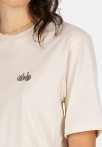 Cream-colored cotton t-shirt with short sleeves, featuring black embroidered bicycle with daisies on the left chest. Soft texture, relaxed fit.