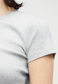 Κοντινό πλάνο ενός ατόμου που φορά γκρι ανοιχτόχρωμο, κοντομάνικο μπλουζάκι με στρογγυλή λαιμόκοψη, εμφανίζοντας την περιοχή του ώμου και του άνω βραχίονα.