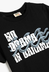 Sort bomuld t-shirt med hvid og lyseblå retro tekstdesign med bølgede accenter, der står "NO DRAMA I BAHAMAS." Rund hals.