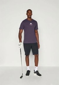 Άντρας με σκούρο μωβ T-shirt της Nike, μαύρες βερμούδες και αθλητικά παπούτσια που στέκεται κρατώντας μια ράβδο γκολφ με ένα λευκό γάντι στο αριστερό του χέρι.