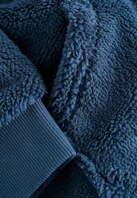 Námornícky modrý fleece materiál s plyšovým, textúrovaným povrchom a ribbovanými pletenými manžetami. Materiál vyzerá mäkko a hrubo, poskytujúc teplo.