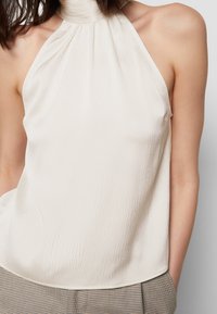 Haut à col halter en tissu texturé crème, avec des plis verticaux et une silhouette ajustée. Associé à un pantalon taille haute à carreaux.