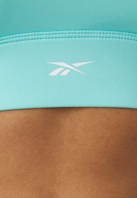 Ανοιχτό τουρκουάζ σπορ σουτιέν με λεία υφή, που διαθέτει λευκό λογότυπο και ραφές κατά μήκος της ζώνης, κοντινή λήψη στην μεσαία περιοχή.