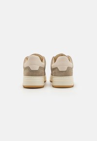 Beige mokkasinsiltä näyttävät tennarit, joissa on vaaleanbeige nahka aksenttina ja tekstuurilla varustetut kantapäät. Pohja on valkoinen ja siinä on kuminen ulkopohja. Minimalistinen muotoilu.