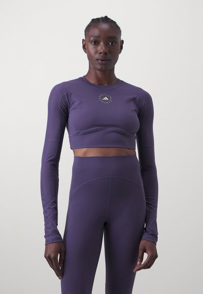 Fialový dlhý rukáv crop top s okrúhlym výstrihom, na ktorom sa nachádza zelené logo. Materiál je hladký a priliehavý, skombinovaný s legínami s vysokým pásom.