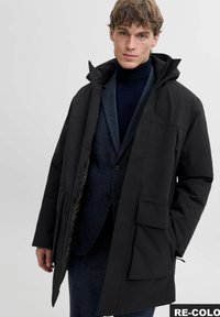 Čierna nepremokavá parka so zipsom vpredu, veľkými vreckami a kapucňou, nosená cez modrý štruktúrovaný blazer s faux golierom. Hladký povrch látky.