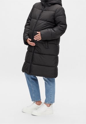 Seitenansicht einer Person, die einen schwarzen Steppmantel, blaue Jeans und weiße Sneaker trägt und ihren Babybauch hält.