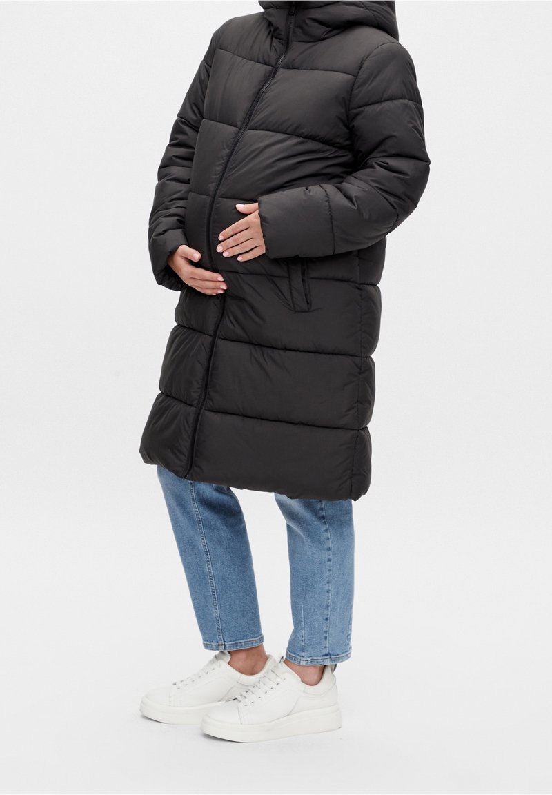 Vue de profil d'une personne portant un manteau matelassé noir, un jean bleu et des baskets blanches, tenant son ventre de femme enceinte.