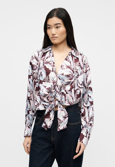 Γυναίκα με μακριά σκούρα μαλλιά που φοράει δέσιμο σε φλοράλ μπλούζα σε λευκούς, ροζ και μοβ τόνους και σκούρο μπλε τζιν, στέκεται μπροστά σε μονόχρωμο φόντο.