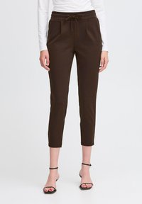 Naine kannab lühikesi tumesiniseid paeltega pükse, millel on ees taskud, ja musti rihmadega kontsasandaale, seistes valge tausta ees.