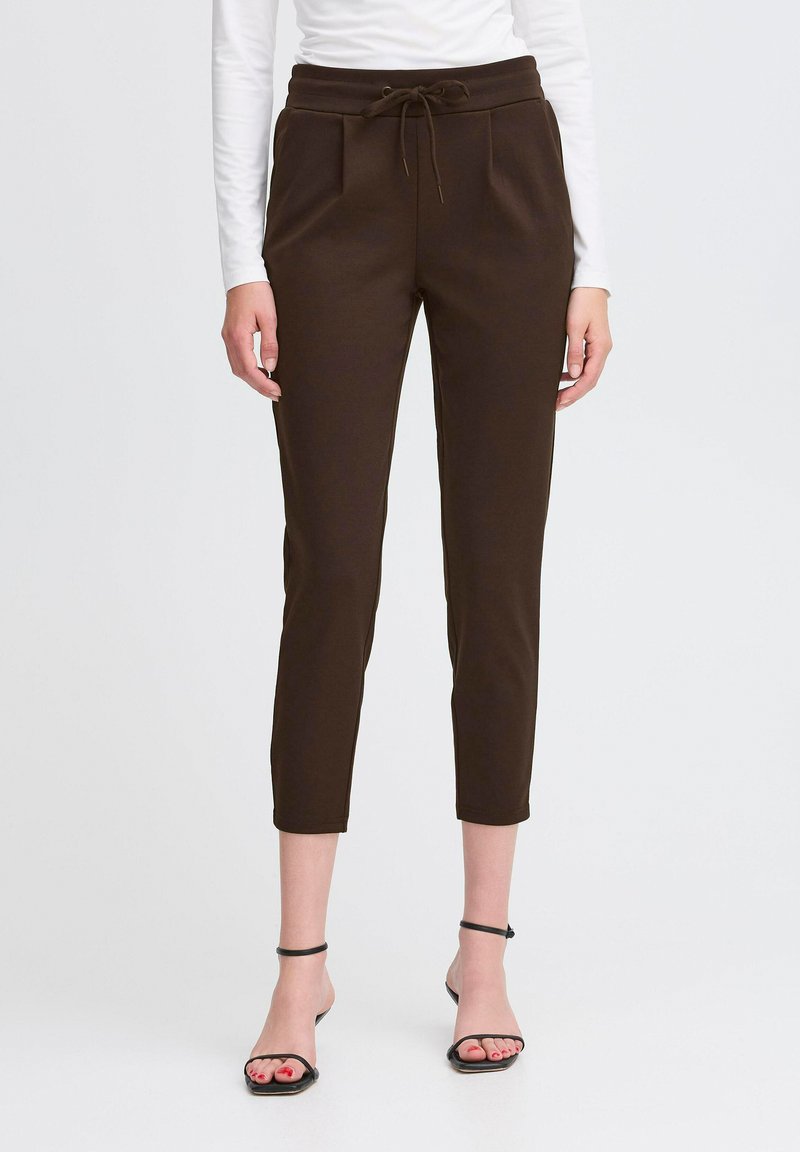 Naine kannab lühikesi tumesiniseid paeltega pükse, millel on ees taskud, ja musti rihmadega kontsasandaale, seistes valge tausta ees.