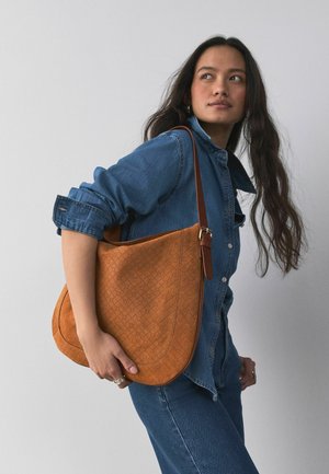 Mujer con chaqueta vaquera y vaqueros sostiene un gran bolso de hombro de cuero marrón texturizado, mirando hacia su izquierda sobre un fondo gris liso.