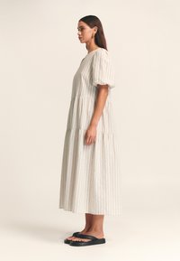 Robe maxi légère à rayures avec manches bouffantes, design à volants et texture douce. Portée avec des sandales plates noires. Couleur beige neutre.