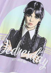 T-shirt violet avec un personnage illustré ayant des cheveux noirs en tresses, portant une tenue noire et un col blanc, avec le texte "Mercredi".