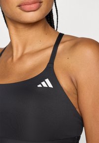 Μαύρο αθλητικό σουτιέν με λεπτές τιράντες, με λευκό λογότυπο τριών ραβδώσεων στην αριστερή πλευρά, κατασκευασμένο από απαλό, ελαστικό υλικό.